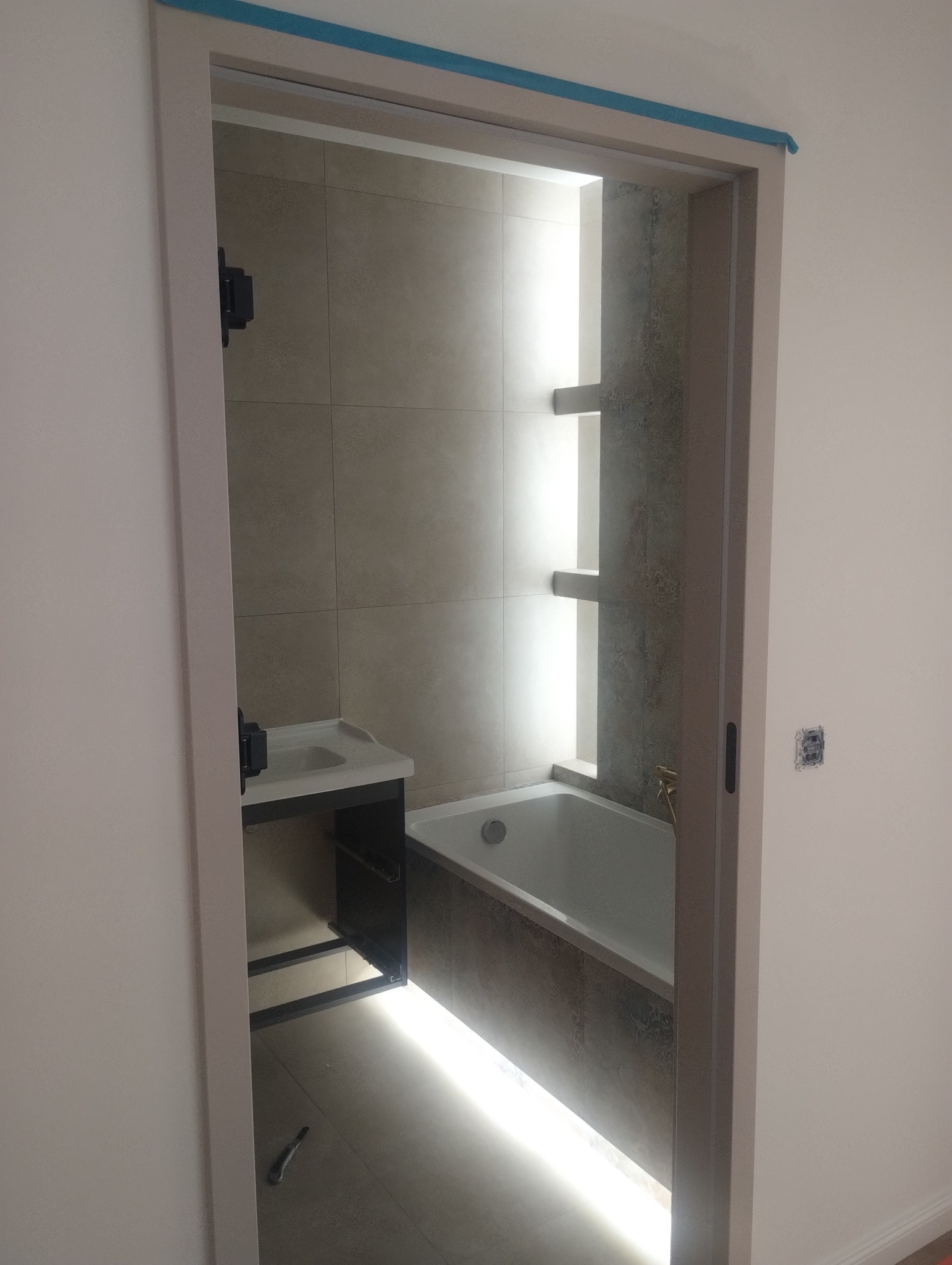 Wykończona łazienka z wanną, umywalką i podświetleniem LED, widoczna przez futrynę drzwiową. Minimalistyczny design z szarymi płytkami i akcentami światła.