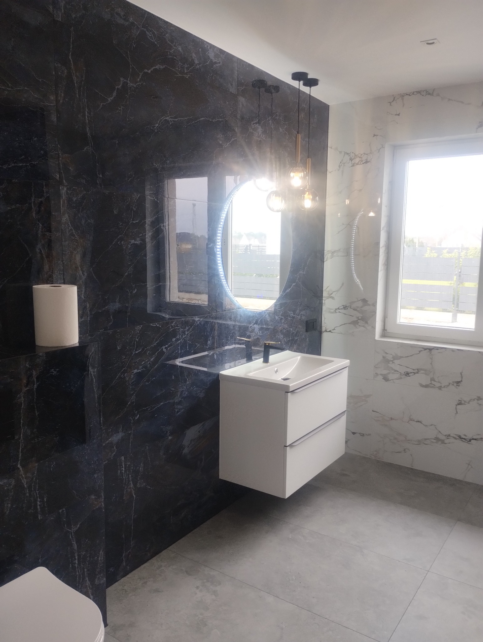 Nowoczesna łazienka z czarnymi i białymi marmurowymi płytkami, okrągłym lustrem z podświetleniem LED i wiszącą szafką pod umywalkę. Designerskie oświetlenie nad lustrem.