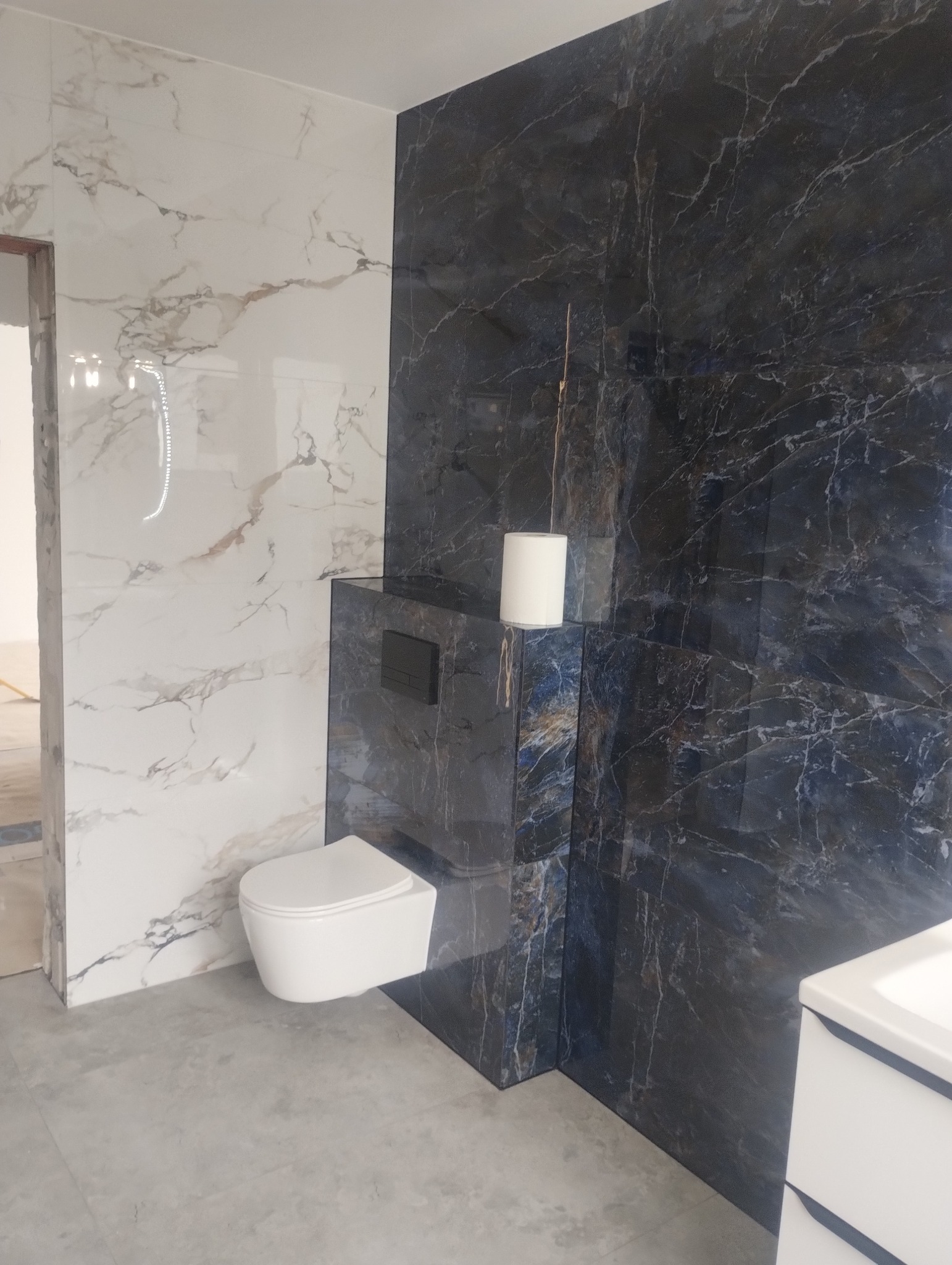 Wykończona łazienka z marmurowymi płytkami w odcieniach bieli i granatu, wisząca toaleta i zabudowa podtynkowa. Minimalistyczny design z akcentem na naturalne wzory.