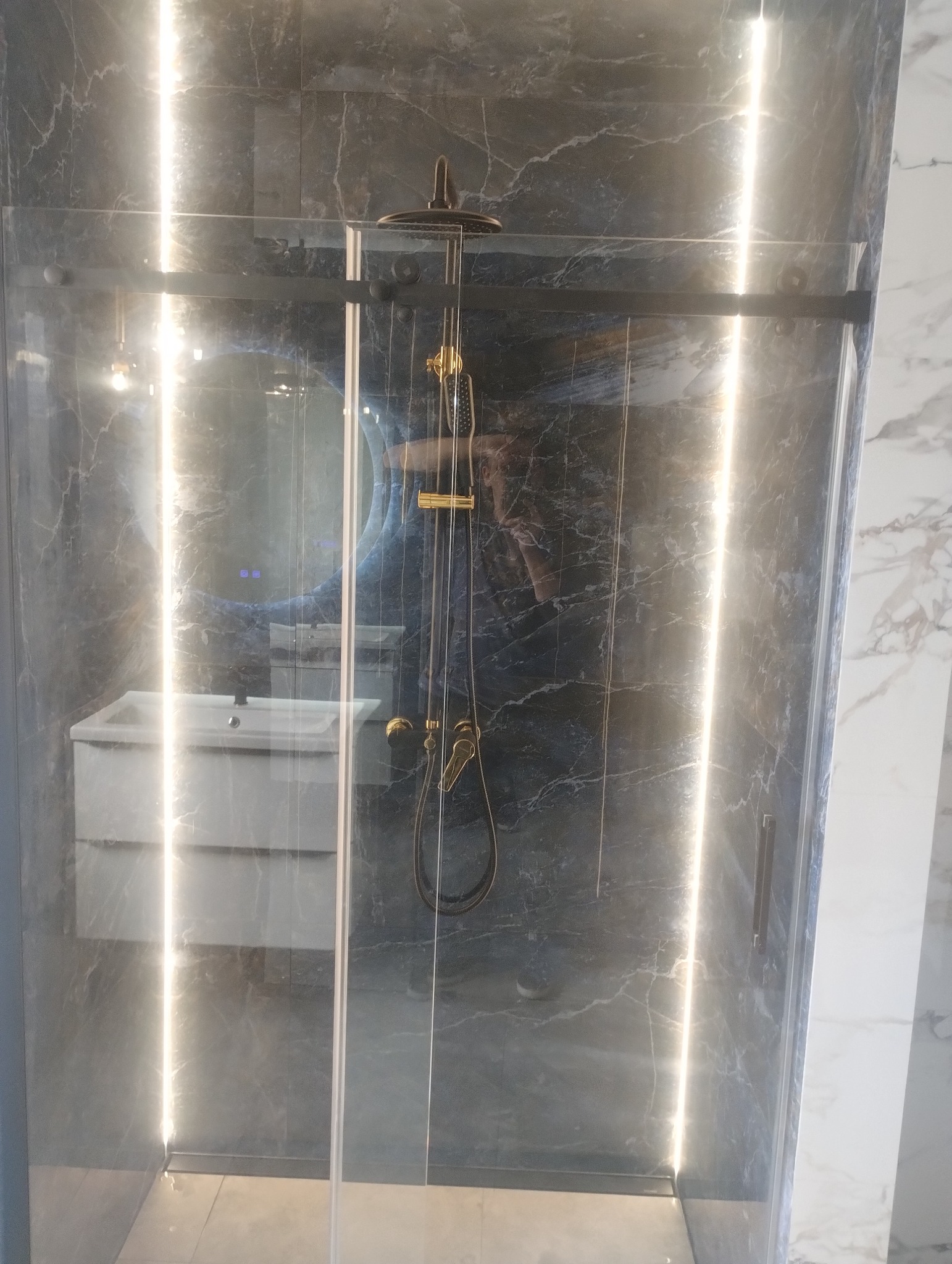 Szklana kabina prysznicowa z deszczownicą w łazience wykończonej marmurem. Widoczne oświetlenie LED i umywalka z szafką.