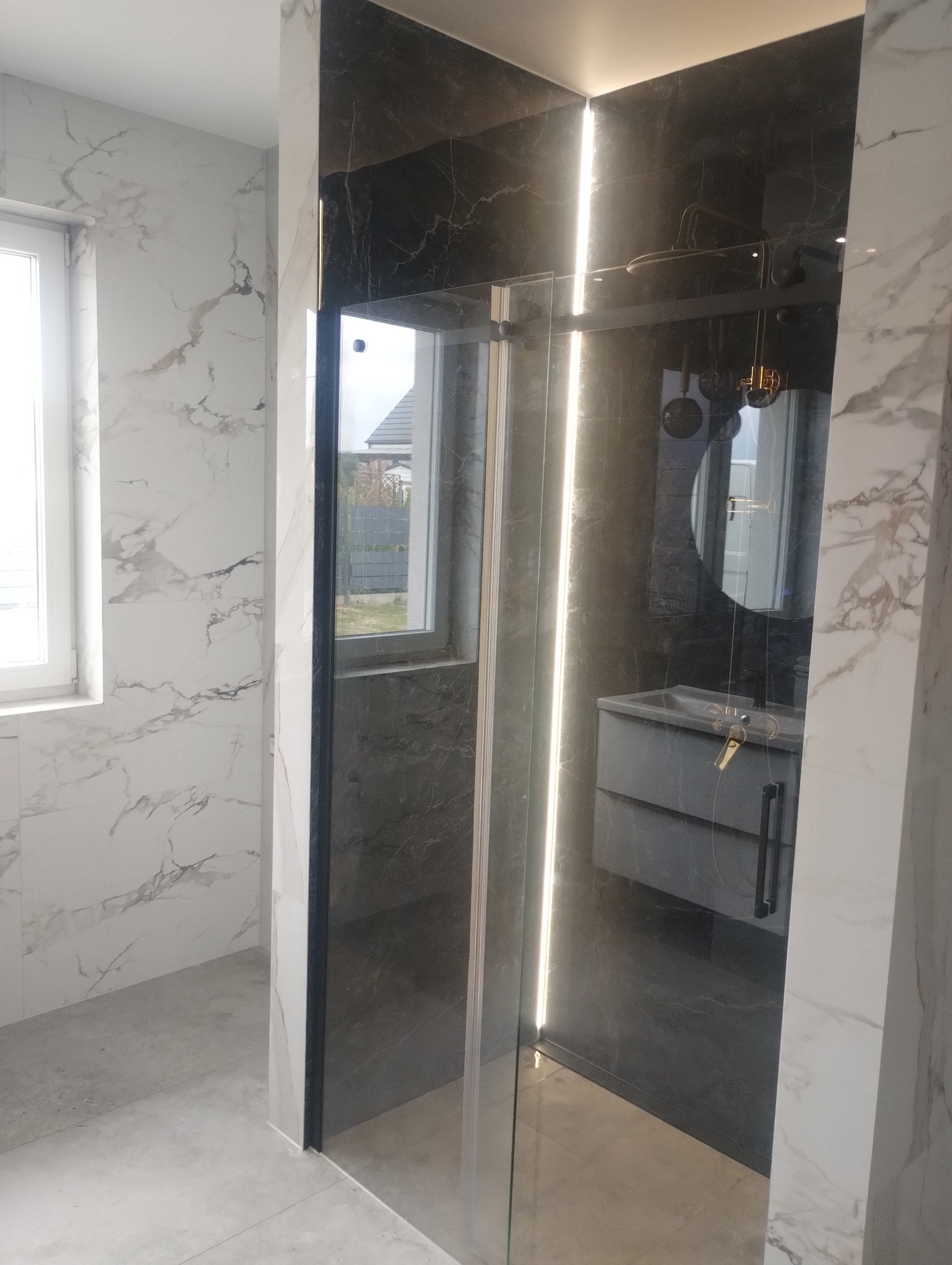Nowoczesna łazienka z kabiną prysznicową typu walk-in, wykończona płytkami imitującymi marmur w odcieniach bieli i szarości, z subtelnym oświetleniem LED.