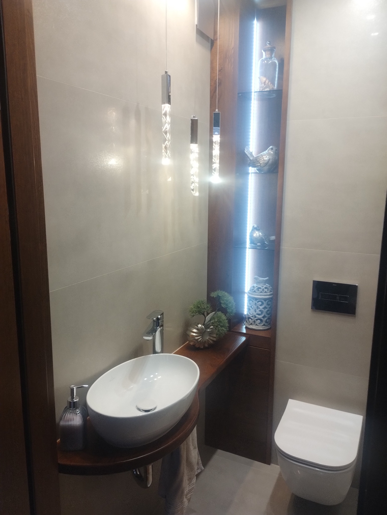 Nowoczesna łazienka z owalną umywalką na drewnianym blacie, podwieszaną toaletą i podświetlaną półką z dekoracjami. Wiszące lampy kryształowe.