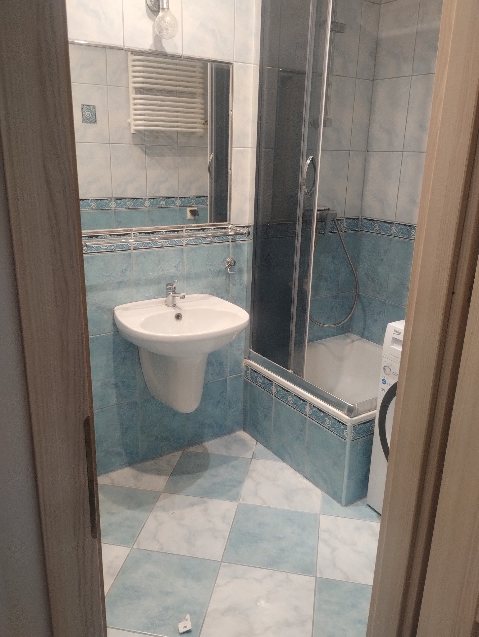 Jasna łazienka po remoncie z umywalką, kabiną prysznicową i pralką. Błękitne płytki na ścianach i podłodze. Widok zza otwartych drzwi.