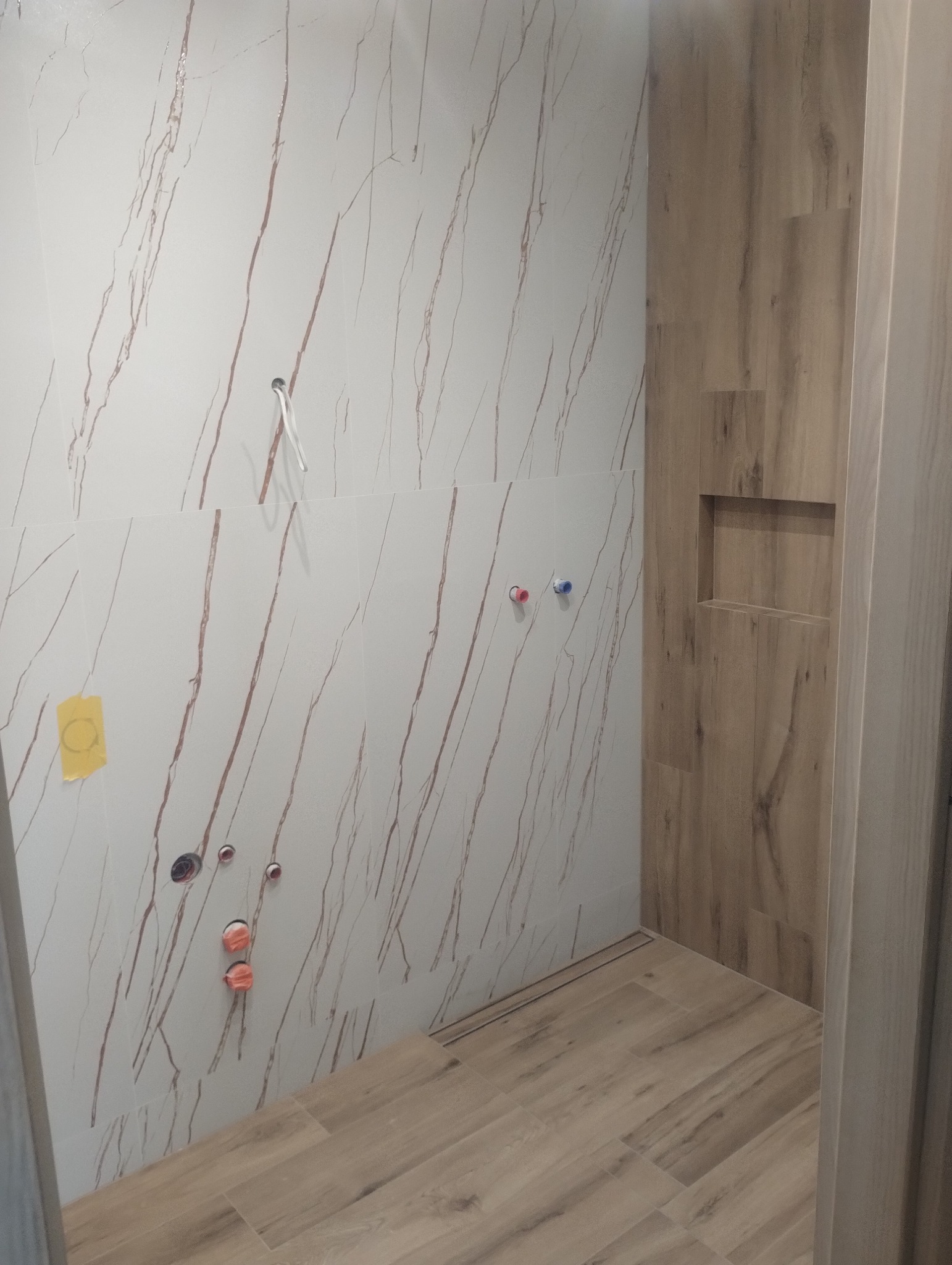 Wykończona łazienka w beżach: płytki imitujące drewno na podłodze i ścianie wnęki, marmurowe płytki z brązowymi żyłami na pozostałych ścianach. Widoczne przyłącza hydrauliczne.