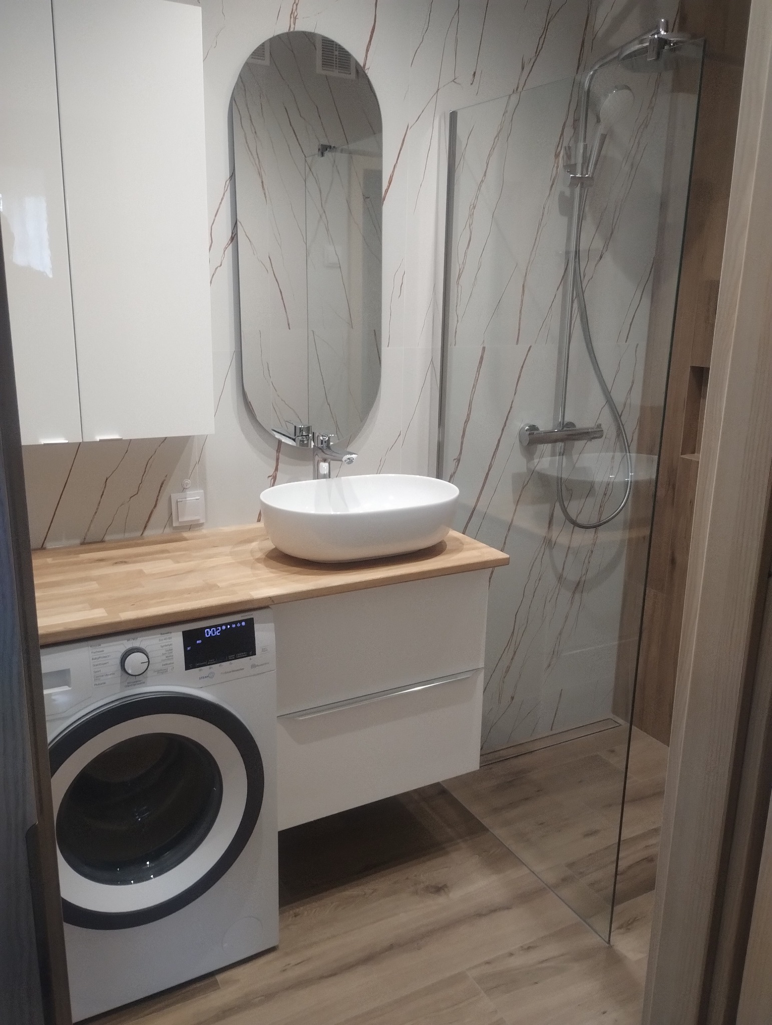 Nowoczesna łazienka z kabiną prysznicową, umywalką nablatową i pralką. Białe szafki i drewniany blat dodają elegancji. Ściany wykończone płytkami imitującymi marmur.