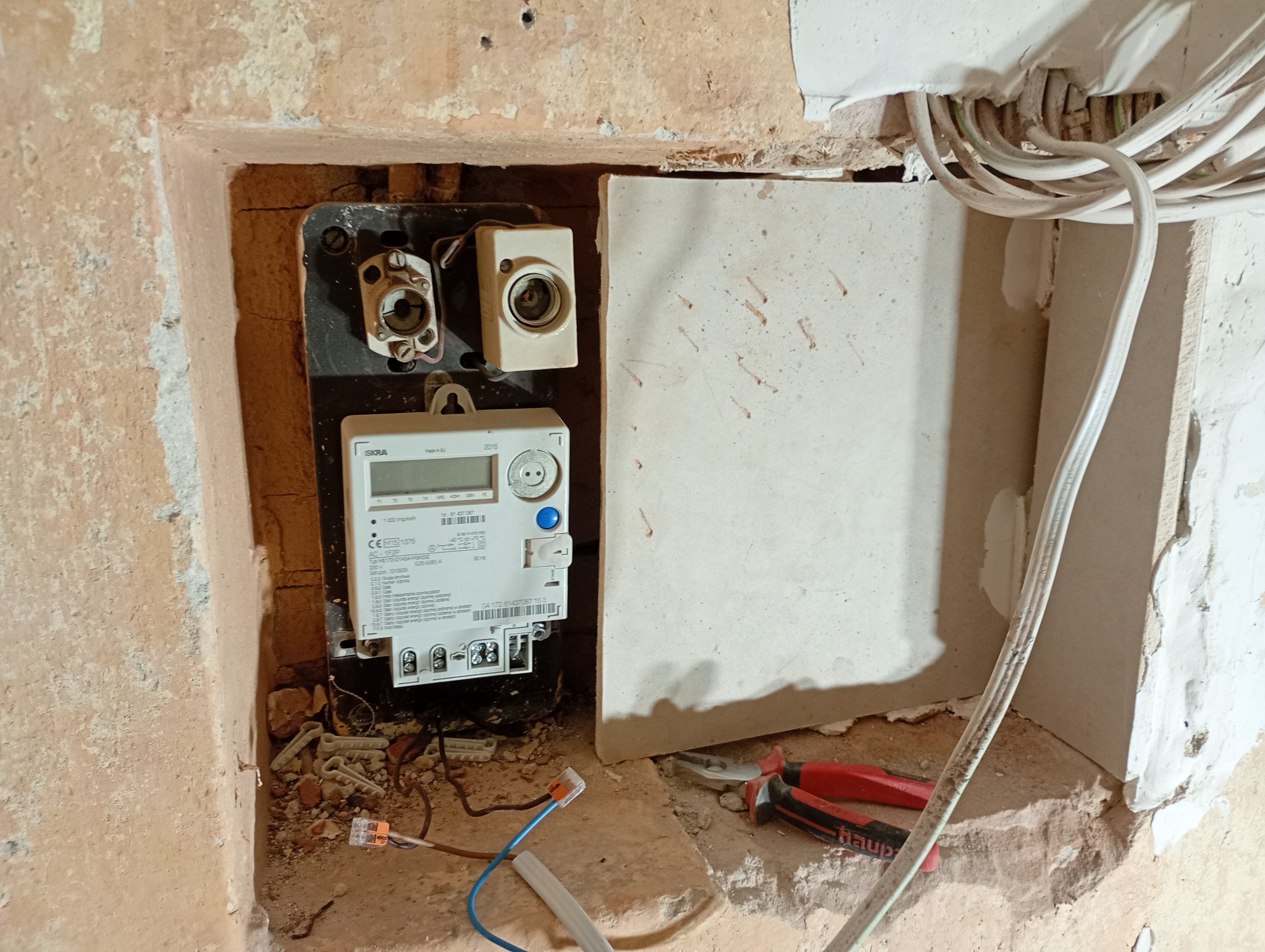 Wnęka z licznikiem elektrycznym i przewodami w trakcie prac instalacyjnych. Widoczne narzędzia i elementy instalacji. Ściany w stanie surowym, przed wykończeniem.