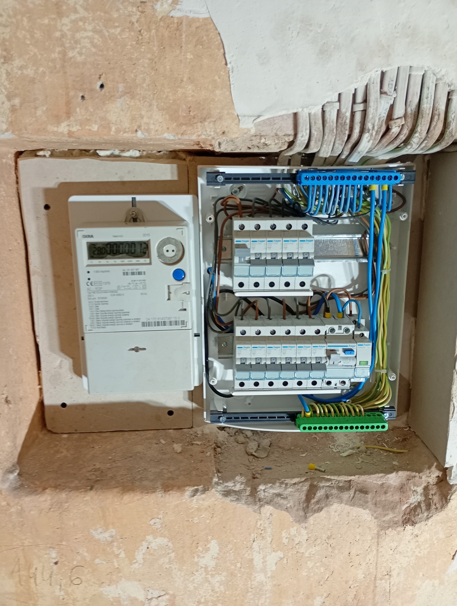 Szczegółowe ujęcie na zamontowany licznik energii elektrycznej obok skrzynki z bezpiecznikami, umieszczone w surowym otworze w ścianie z widocznymi przewodami.