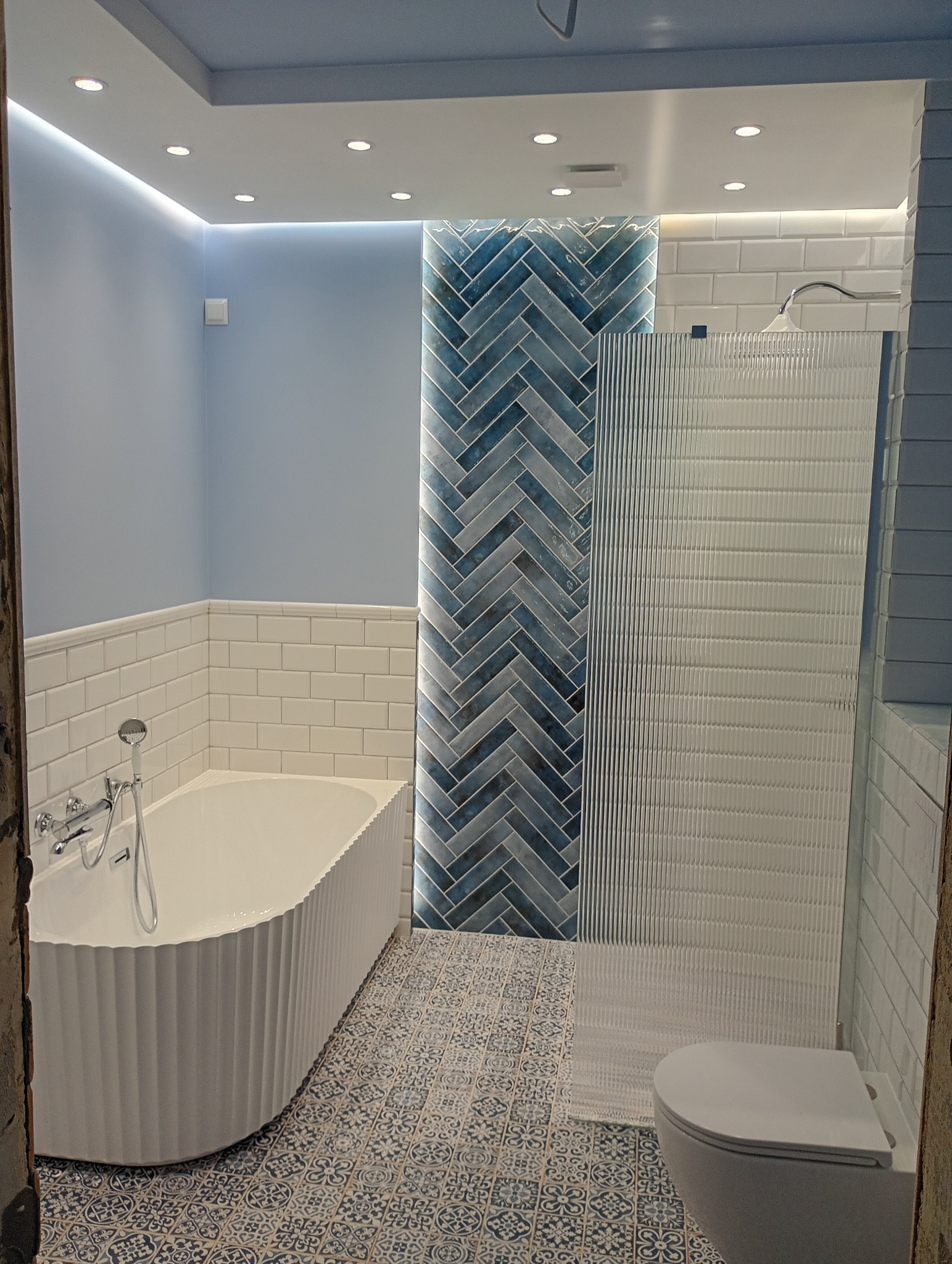 Wykończona łazienka w Stargardzie: biała wanna z dekoracyjną obudową, ściana z niebieskiej jodełki i kabina prysznicowa z ryflowanego szkła. Podłoga w dekoracyjne wzory.