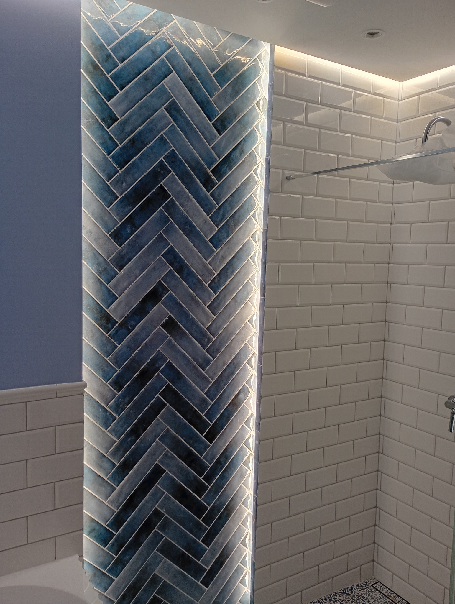 Ściana w łazience z niebieską mozaiką ułożoną w jodełkę, podświetloną listwą LED. Obok białe, prostokątne płytki i fragment kabiny prysznicowej.