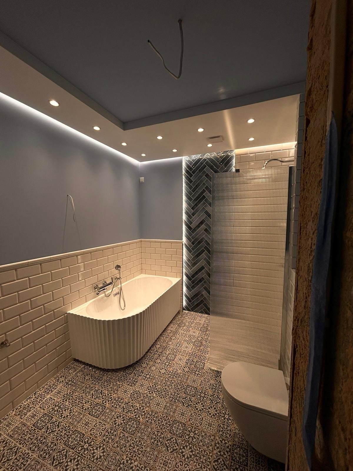 Nowoczesna łazienka z wanną, toaletą i kabiną prysznicową. Uwagę zwracają dekoracyjne płytki na podłodze i ścianie, oraz oświetlenie LED pod sufitem.