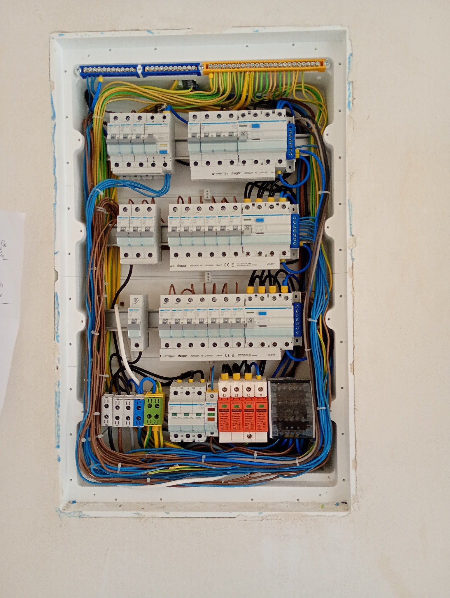 Rozdzielnica elektryczna Hager w białej obudowie, z widocznymi bezpiecznikami, przewodami w różnych kolorach i listwami zaciskowymi, umieszczona w ścianie w trakcie montażu.