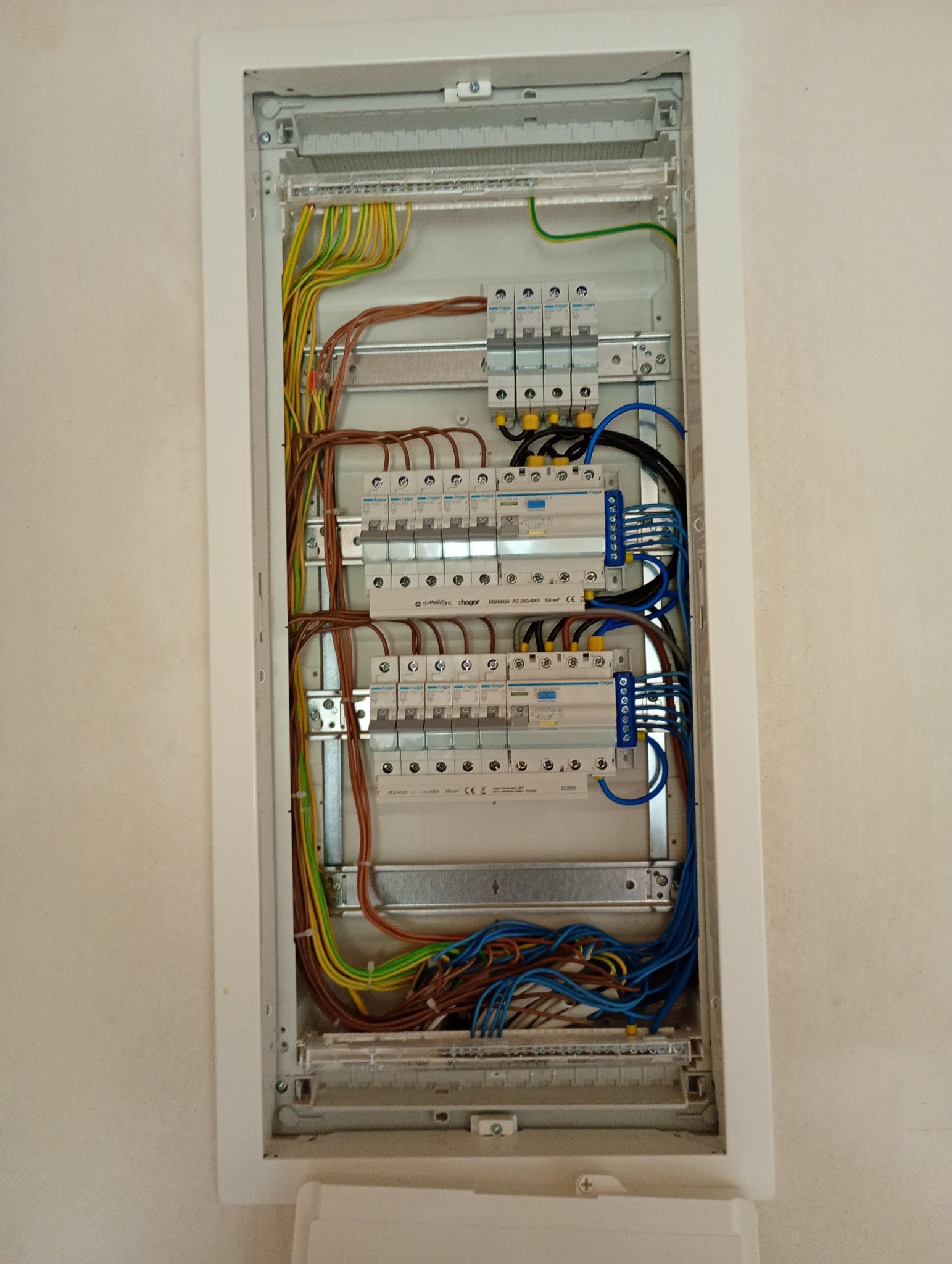 Szczegółowe ujęcie rozdzielnicy elektrycznej w białej obudowie, z widocznymi kablami w kolorach: niebieskim, brązowym, żółto-zielonym, oraz zabezpieczeniami.
