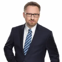 Portret mężczyzny w okularach, granatowym garniturze i krawacie w niebieskie paski, na białym tle.