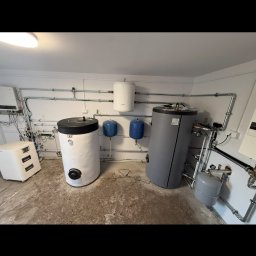 TOM-BUD - Kompletna instalacja CO w kotłowni: dwa zbiorniki akumulacyjne, bojler, naczynia wzbiorcze i orurowanie. Białe ściany, betonowa posadzka. Widok z perspektywy podłogi.