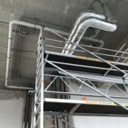 Wnętrze w trakcie prac instalacyjnych: metalowe rury wentylacyjne w izolacji, rury instalacji hydraulicznej pod sufitem oraz rusztowanie z widocznym napisem 'Mietgerüst Rieckermann'.