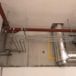 Widok instalacji pod sufitem: rury miedziane, rury w izolacji termicznej i rury PCV w kolorze ceglastym, mocowane do sufitu za pomocą metalowych wsporników i prętów.