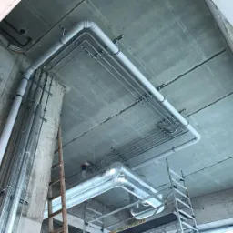 Widok instalacji rur pod sufitem w surowym stanie, z widocznymi elementami mocującymi i rusztowaniem w tle.