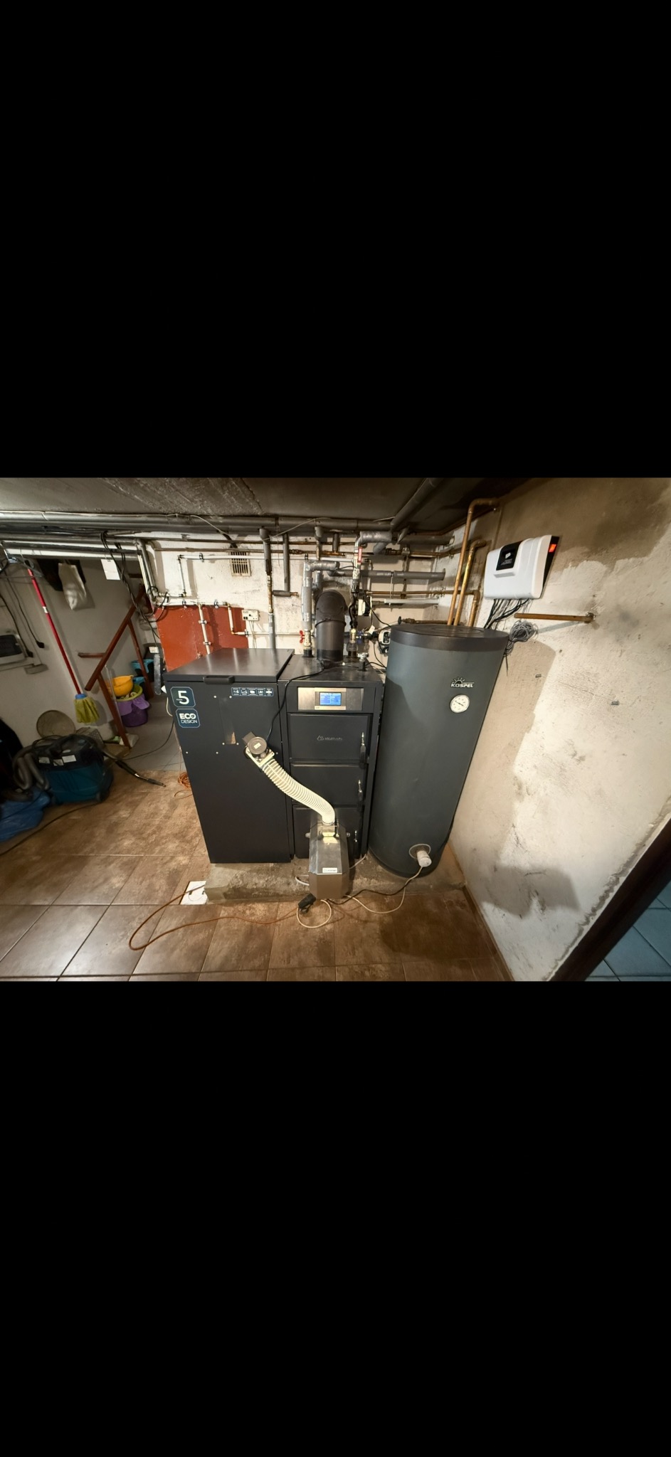 Nowoczesny piec CO z zasobnikiem Kospel w kotłowni, widoczne rury instalacji grzewczej. Całość w pomieszczeniu z płytkami podłogowymi.