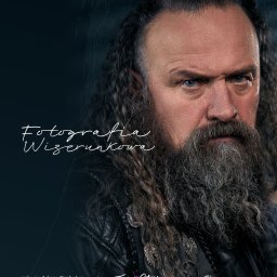 Grupa TWOJE.STUDIO - Portret mężczyzny z długą brodą i włosami, w skórzanej kurtce z przypinkami, na ciemnym tle. Wyraziste spojrzenie, ostre światło. Fotografia wizerunkowa.