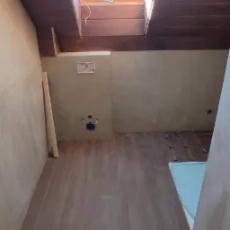 Łazienka w trakcie remontu: widoczny stelaż podtynkowy WC, częściowo położone płytki podłogowe imitujące drewno, okno dachowe w drewnianej obudowie, ściany przygotowane pod gładzie.