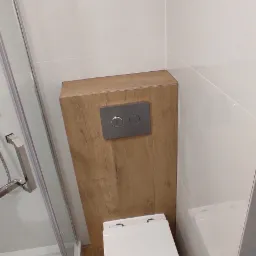 Nowoczesna, minimalistyczna łazienka z podwieszaną toaletą, obudowaną drewnopodobną płytą z chromowanym panelem spłukującym, białe płytki ścienne i podłogowe, kabina prysznicowa z chromowanymi...