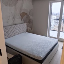 RENOVA - Jasna sypialnia po odświeżeniu: łóżko z tapicerowanym zagłówkiem, wzorzysta tapeta w odcieniach beżu, okno balkonowe z widokiem na osiedle. Minimalistyczny design.