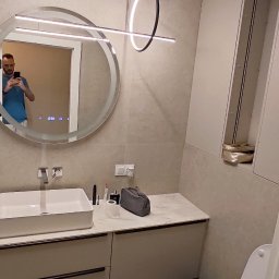 RENOVA - Nowoczesna łazienka z okrągłym lustrem z podświetleniem LED, wiszącą lampą pierścieniową i beżowymi płytkami. W lustrze odbija się osoba robiąca zdjęcie.