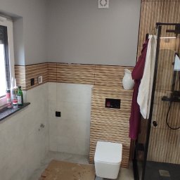 Nowoczesna łazienka w trakcie wykańczania: drewniane panele, biała ceramika, szklana kabina prysznicowa. Podłoga zabezpieczona kartonem.