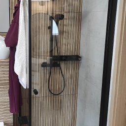 Nowoczesna kabina prysznicowa z czarną armaturą i drewnianą ścianą w tle. Widoczne ręczniki i fragment toalety. Wykończenie łazienki w minimalistycznym stylu.