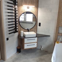 RENOVA - Nowoczesna łazienka z drewnianymi lamelami, okrągłym lustrem z podświetleniem i białą umywalką nablatową. Szare płytki podłogowe i grzejnik drabinkowy.