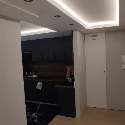 Nowoczesna kuchnia z czarnymi szafkami i blatem z imitacją marmuru, oświetlenie LED na suficie podwieszanym, widok z korytarza na drewnianą podłogę.