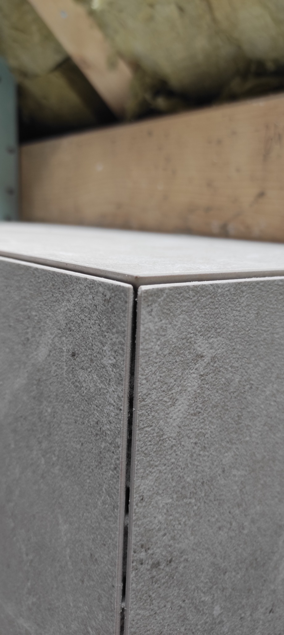 Szczegółowe ujęcie narożnika wykończonego szarą, matową płytką ceramiczną z widoczną fugą, na tle drewnianej konstrukcji i izolacji.