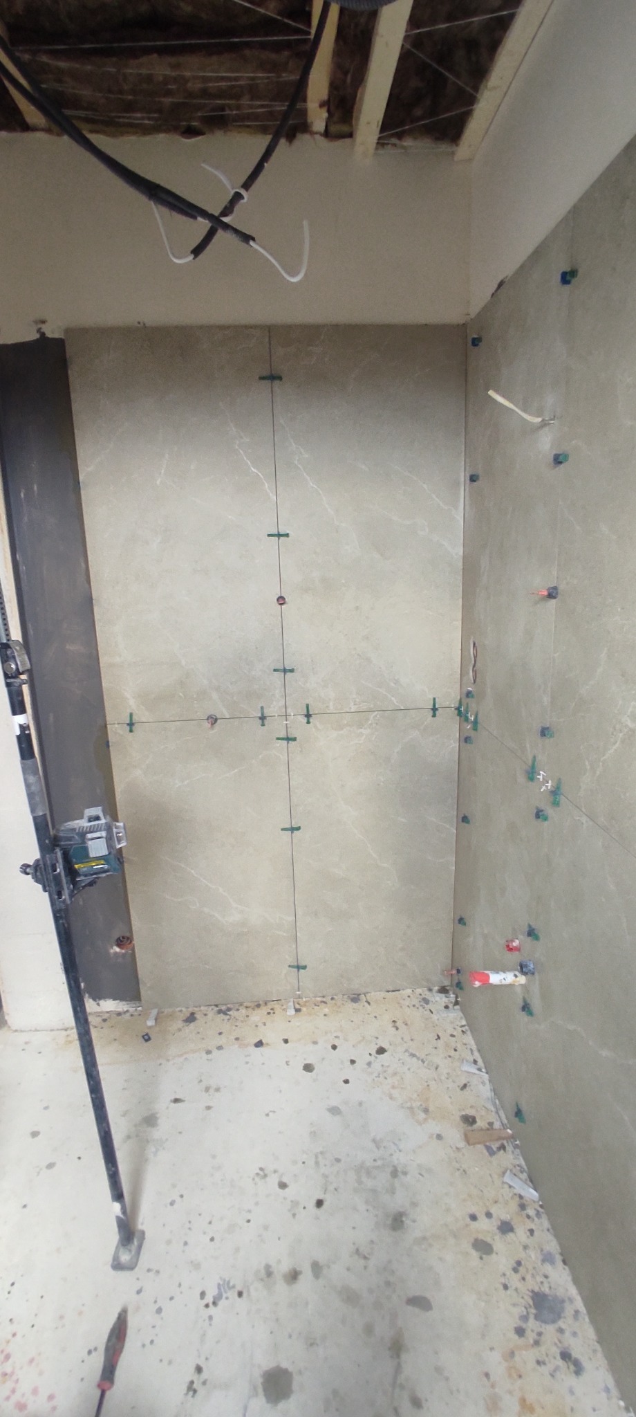 Ściana łazienki w trakcie układania płytek ceramicznych w odcieniach beżu, widoczne krzyżyki dystansowe i poziomica laserowa na statywie, surowy stan podłogi i instalacji sufitowej.