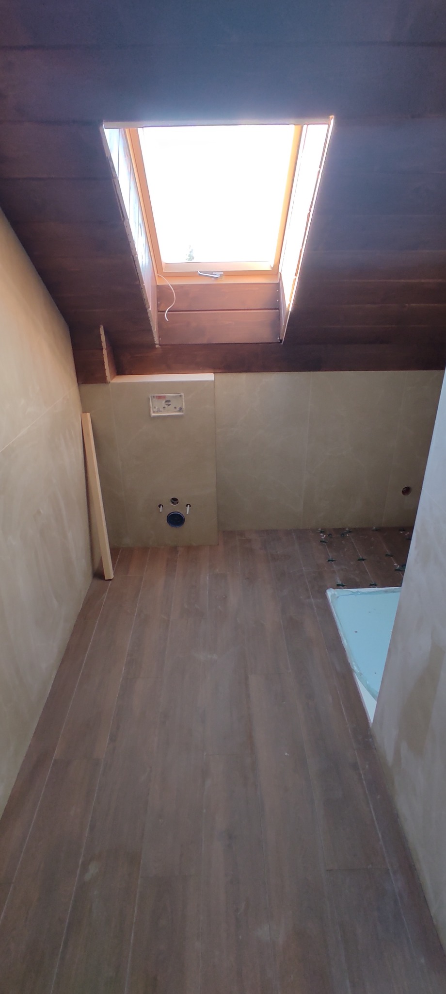 Łazienka w trakcie remontu: widoczny stelaż podtynkowy WC, częściowo położone płytki podłogowe imitujące drewno, okno dachowe w drewnianej obudowie, ściany przygotowane pod gładzie.