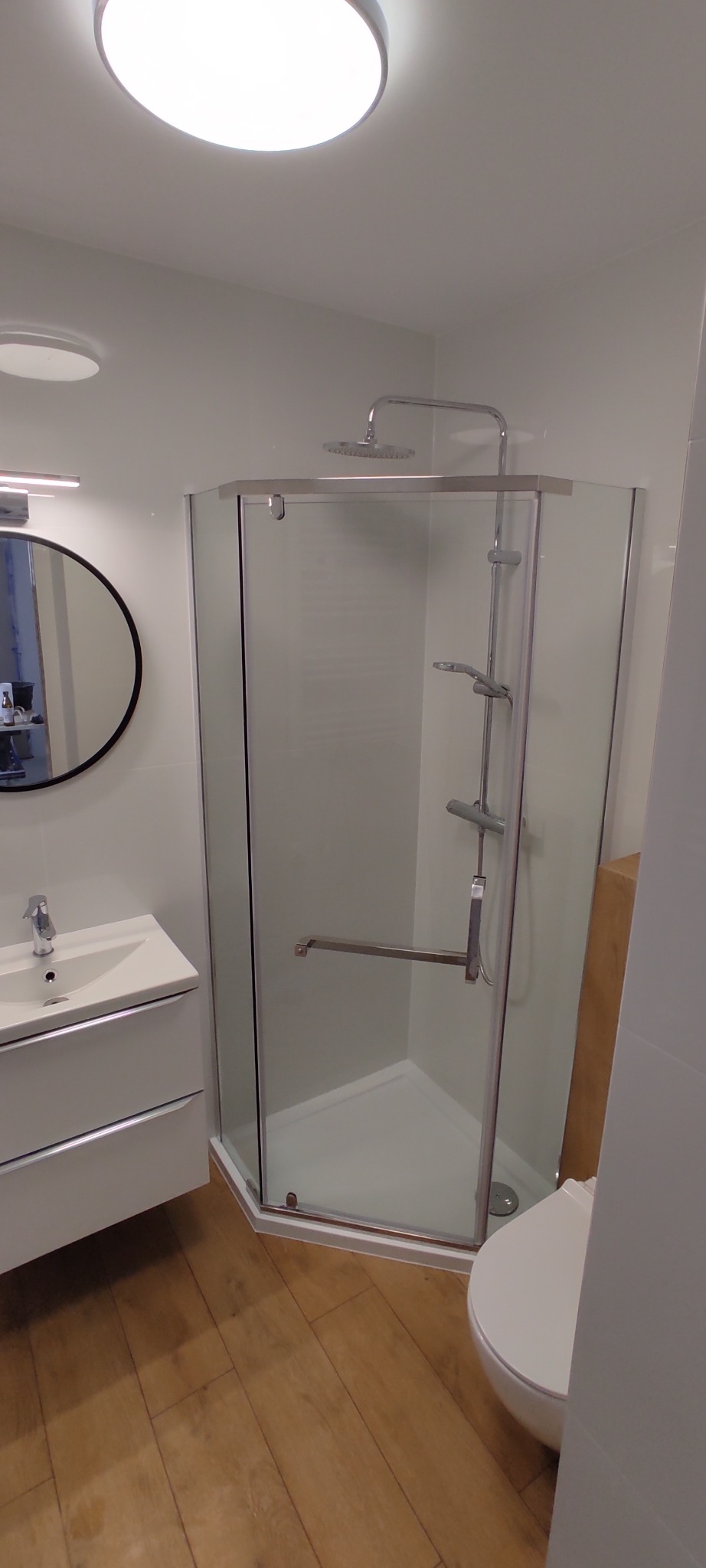 Nowoczesna łazienka z kabiną prysznicową typu walk-in, umywalką nablatową z szafką, okrągłym lustrem i drewnopodobną podłogą.