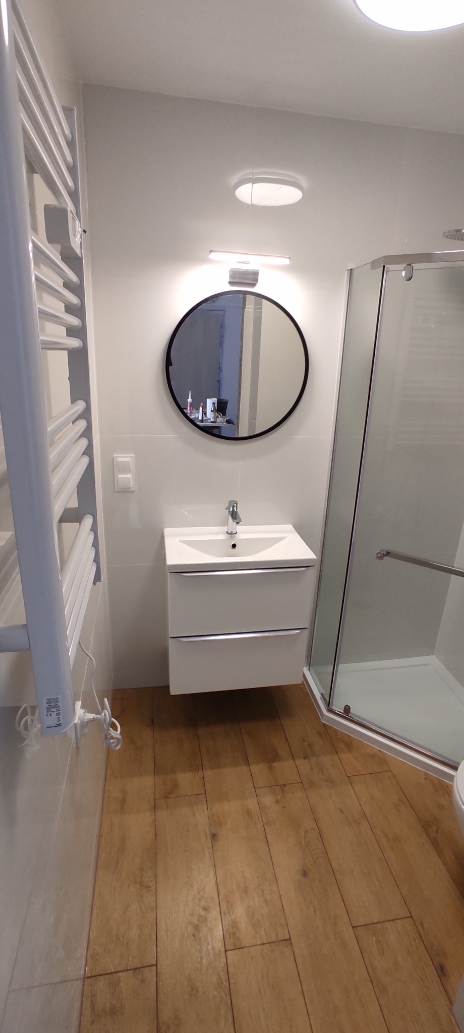 Nowoczesna łazienka z drewnopodobną podłogą, białą szafką z umywalką, okrągłym lustrem z czarną ramą oraz kabiną prysznicową z przezroczystego szkła.