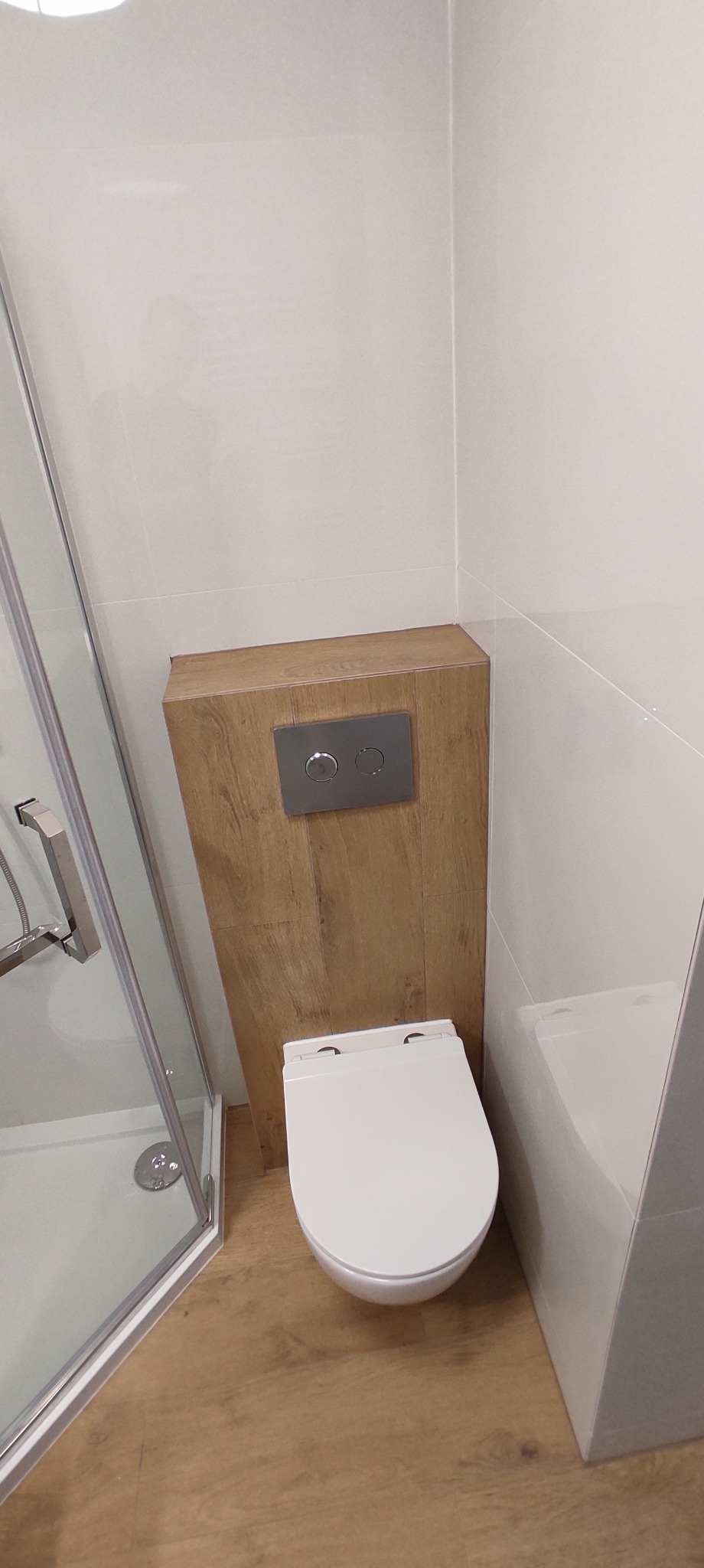 Nowoczesna, minimalistyczna łazienka z podwieszaną toaletą, obudowaną drewnopodobną płytą z chromowanym panelem spłukującym, białe płytki ścienne i podłogowe, kabina prysznicowa z chromowanymi...