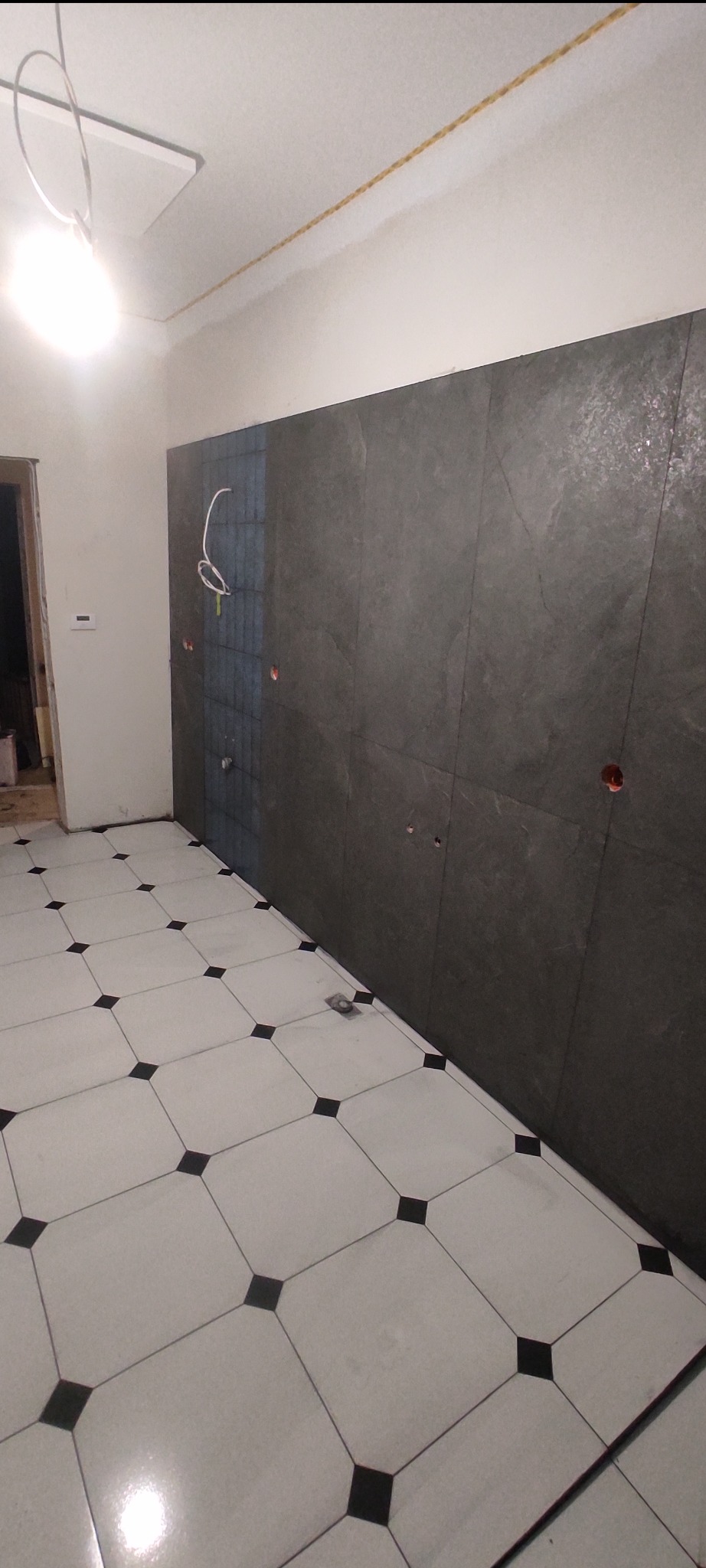 Wykończona łazienka z szarymi płytkami na ścianie i białymi płytkami podłogowymi z czarnymi akcentami. Widoczne przygotowane punkty elektryczne i hydrauliczne.