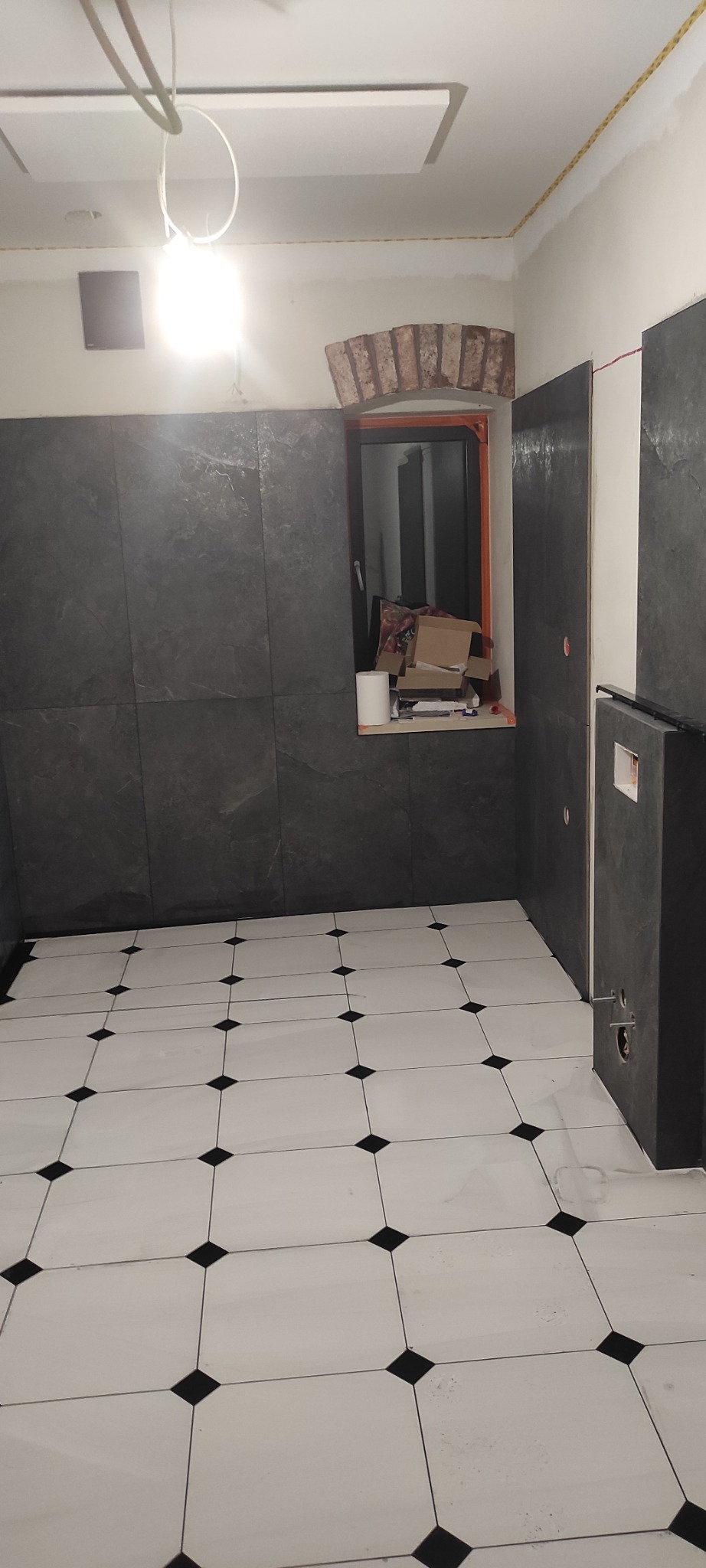 Łazienka w trakcie prac: ciemne płytki ścienne, białe płytki podłogowe z czarnymi akcentami. Widoczne przygotowania do montażu armatury i okno z ceglanym łukiem.