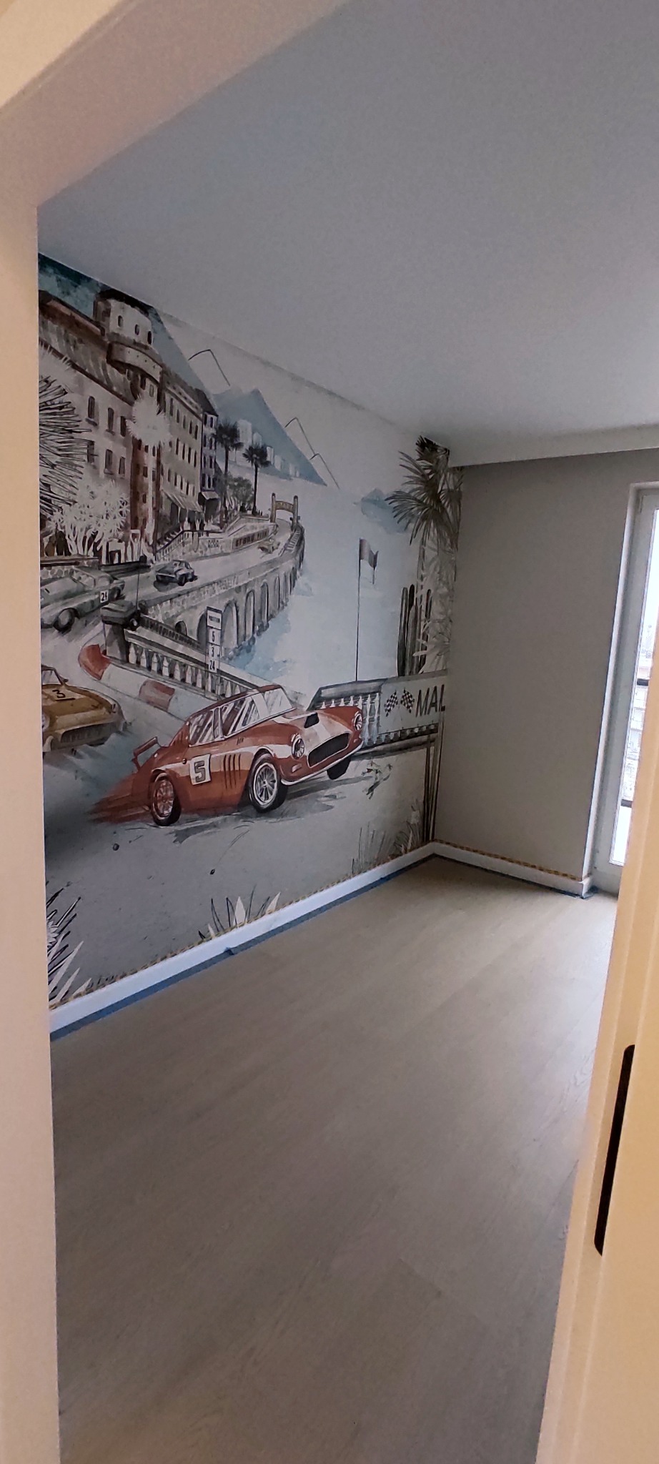 Pokój po malowaniu z fototapetą przedstawiającą wyścigowe samochody i krajobraz miejski. Podłoga wykończona panelami. Widoczne taśmy malarskie przy podłodze.