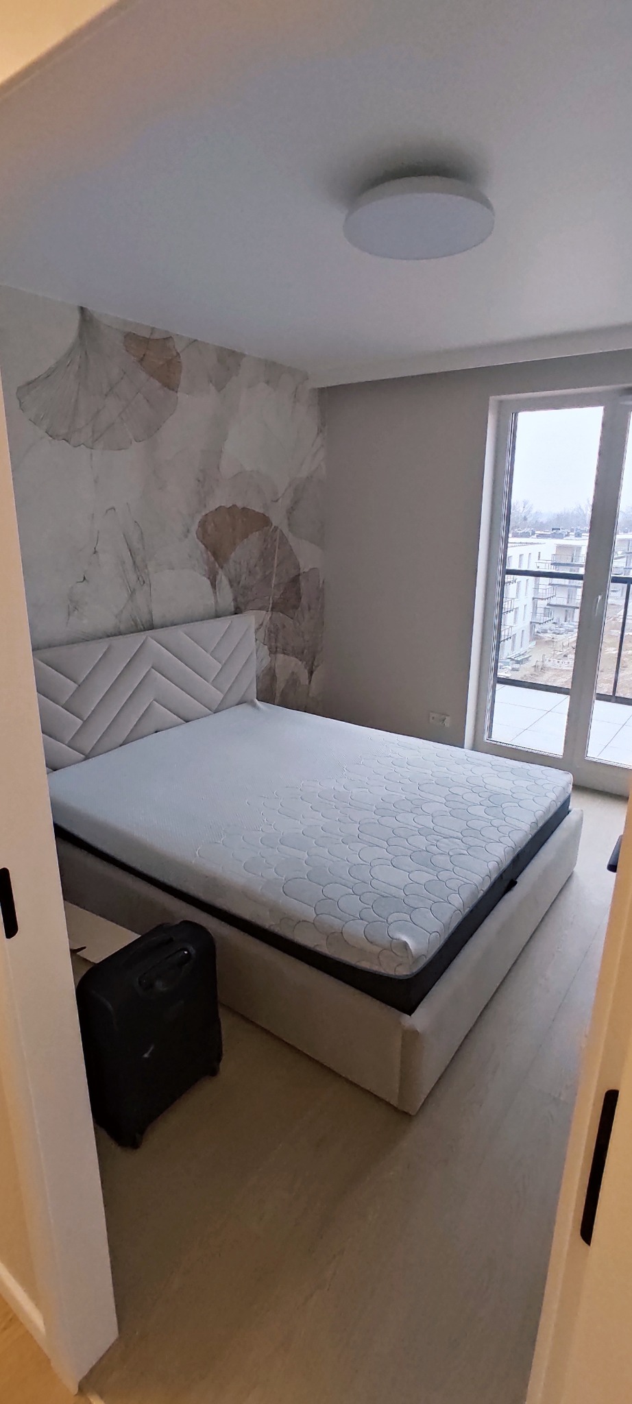 Jasna sypialnia po odświeżeniu: łóżko z tapicerowanym zagłówkiem, wzorzysta tapeta w odcieniach beżu, okno balkonowe z widokiem na osiedle. Minimalistyczny design.