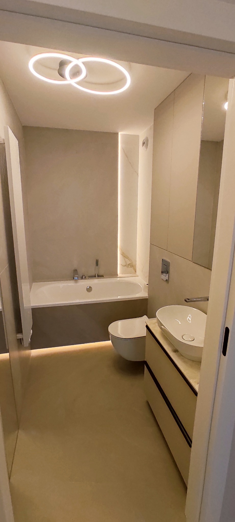 Nowoczesna łazienka w beżowych tonacjach z podświetleniem LED, owalną umywalką nablatową i minimalistyczną armaturą. Widoczna wanna i toaleta.