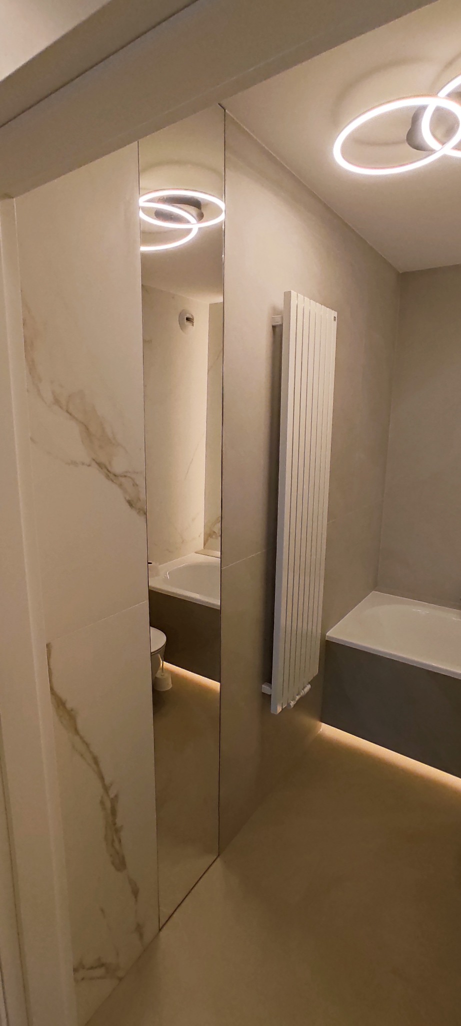 Wykończona łazienka w beżach z podświetleniem LED pod wanną i lustrem. Na ścianach płytki imitujące marmur, grzejnik drabinkowy. Widok z perspektywy wejścia.