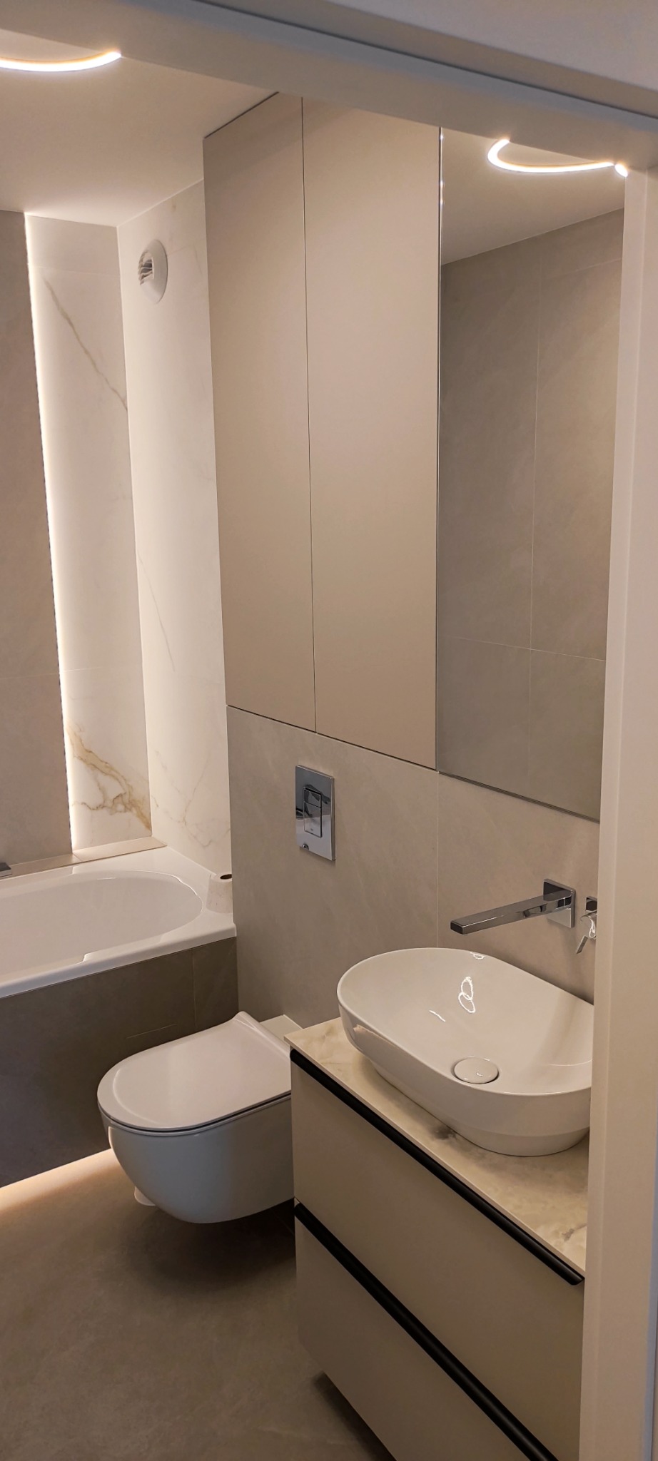 Nowoczesna łazienka w beżach: podwieszana toaleta, umywalka nablatowa, lustro z szafką i subtelne oświetlenie LED. Minimalistyczny design z akcentem na funkcjonalność.