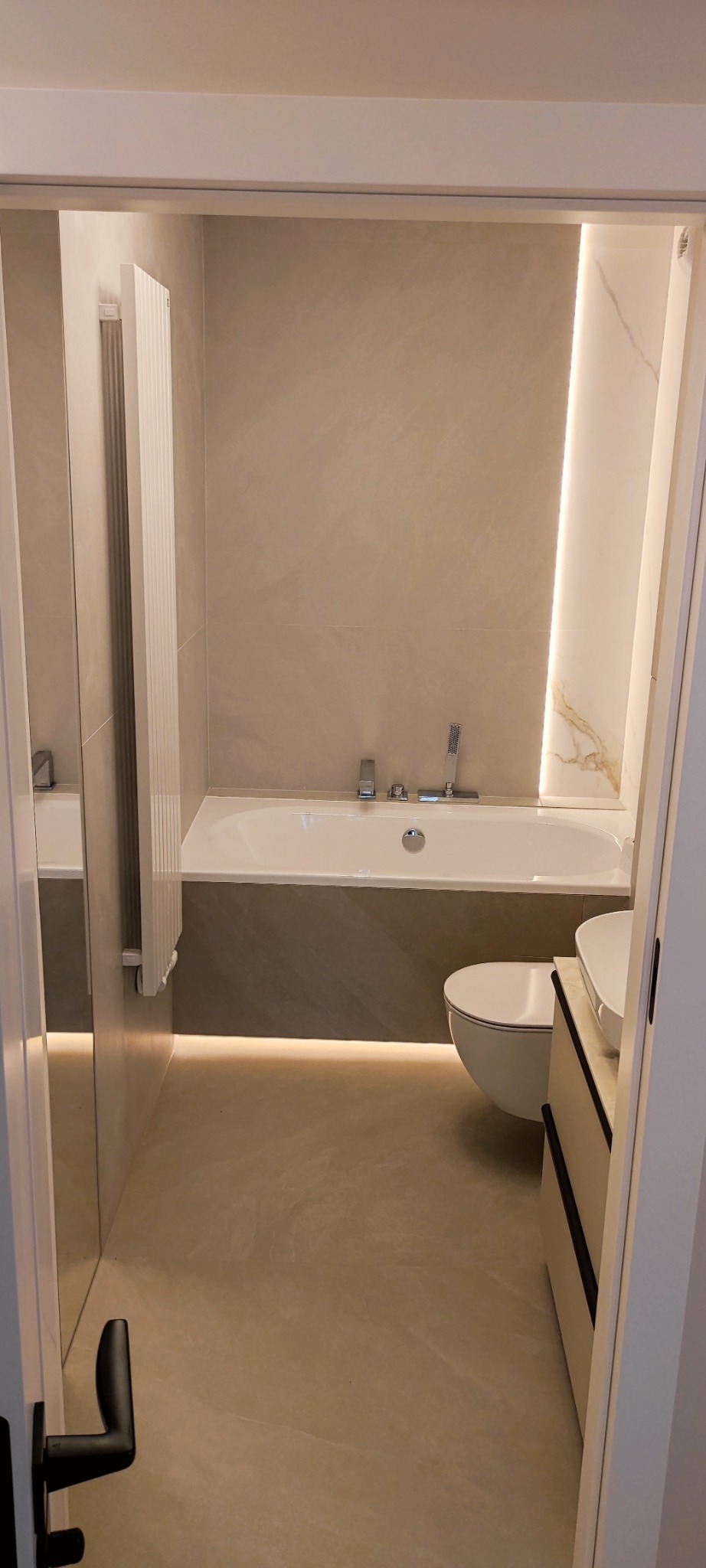 Jasna łazienka z beżowymi płytkami, podświetlaną wanną i umywalką z szafką. Widok zza uchylonych drzwi, podkreślający minimalistyczny design i nowoczesne oświetlenie.