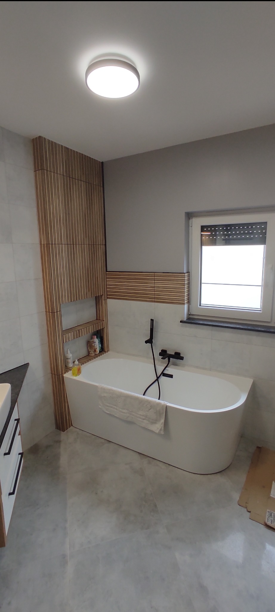 Nowoczesna łazienka z wolnostojącą wanną, drewnianymi panelami ściennymi i szarą marmurową podłogą. Minimalistyczna armatura w czarnym macie.