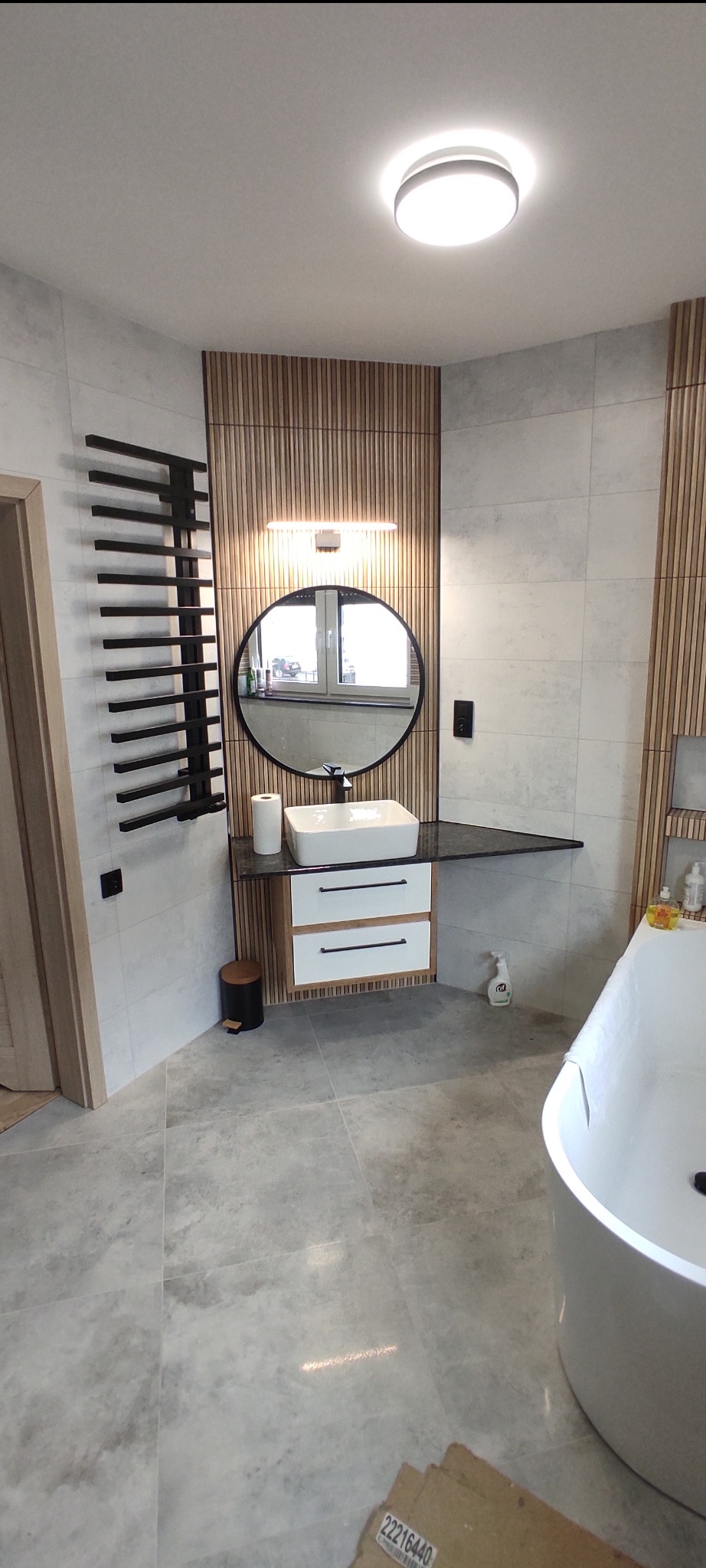 Nowoczesna łazienka z drewnianymi lamelami, okrągłym lustrem z podświetleniem, białą umywalką nablatową i grzejnikiem drabinkowym w kolorze czarnym.