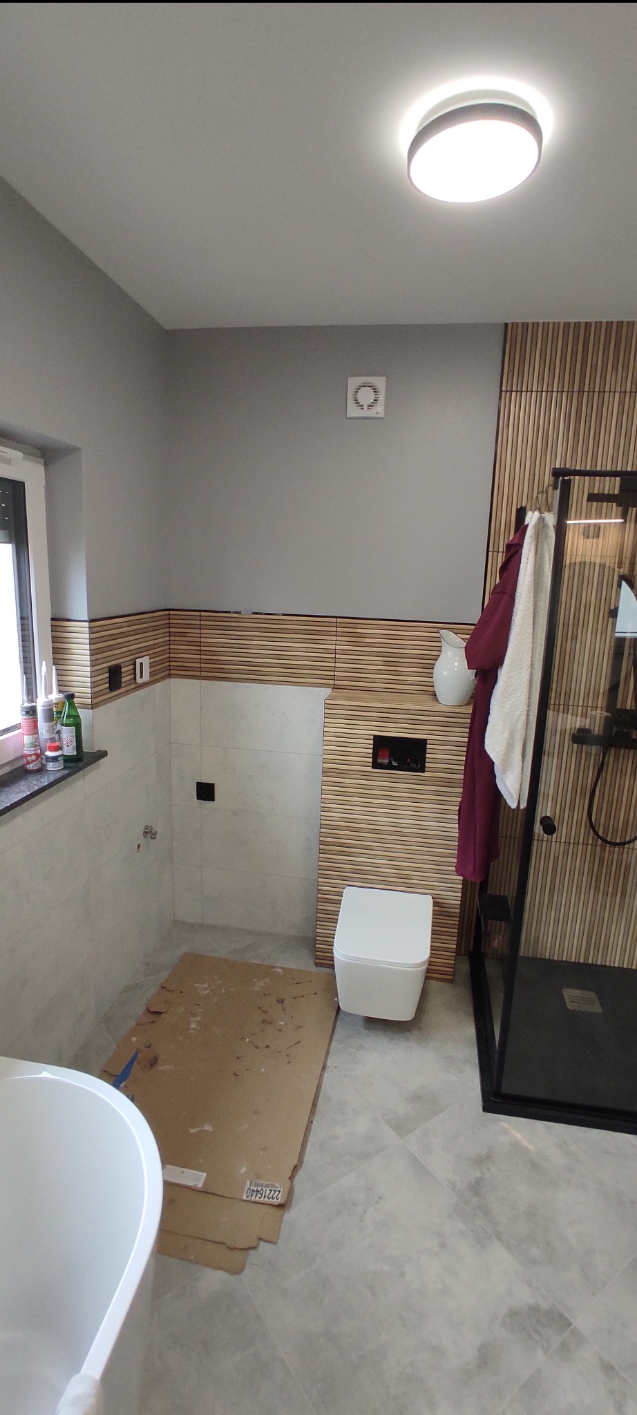 Nowoczesna łazienka w trakcie wykańczania: drewniane panele, biała ceramika, szklana kabina prysznicowa. Podłoga zabezpieczona kartonem.