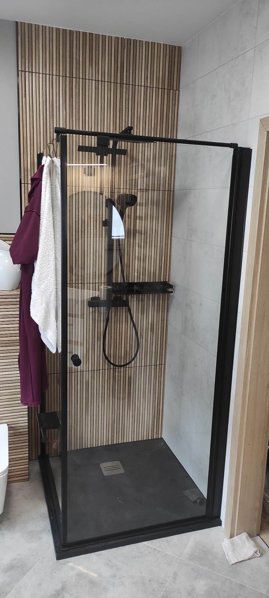 Nowoczesna kabina prysznicowa z czarną armaturą i drewnianą ścianą w tle. Widoczne ręczniki i fragment toalety. Wykończenie łazienki w minimalistycznym stylu.