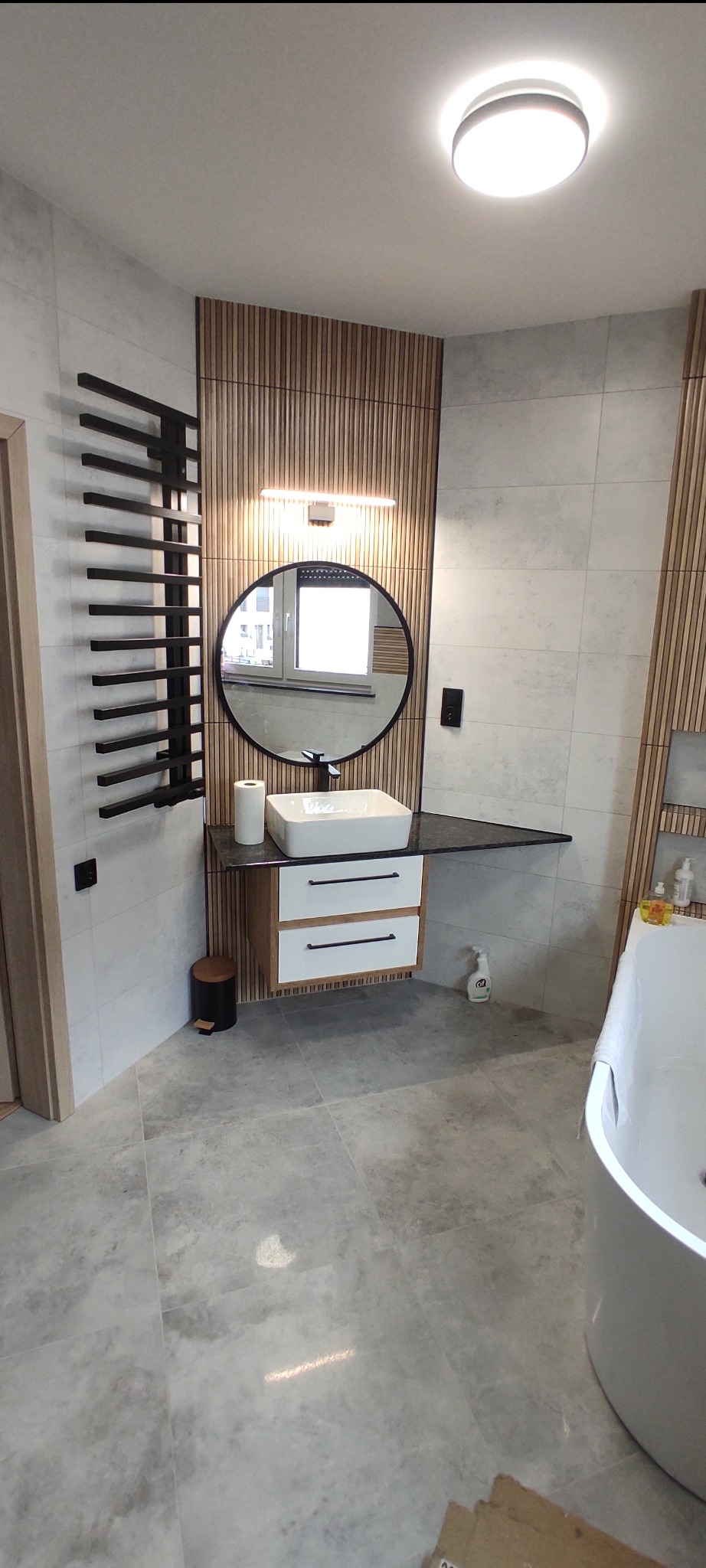 Nowoczesna łazienka z drewnianymi lamelami, okrągłym lustrem z podświetleniem i białą umywalką nablatową. Szare płytki podłogowe i grzejnik drabinkowy.